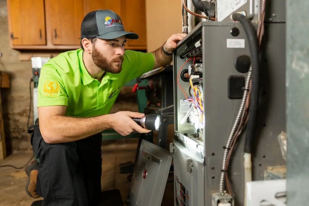 A technician in a green shirt and cap examines the HVAC furnace with a flashlight, inspecting wiring and components in the basement.