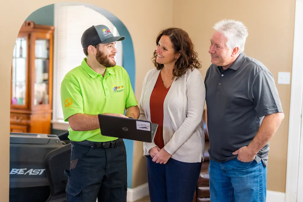 A technician in a green shirt shows a laptop screen to a smiling couple in casual clothes at the St. Charles Home Show, standing in a room with beige walls and wooden furniture.