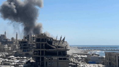 Explosion and smoke rise from a damaged building on Gaza beach amid ongoing conflict.