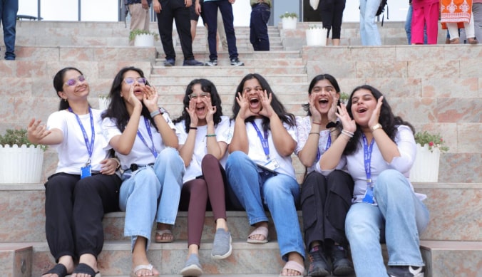 Six women sitting on steps, wearing white shirts and lanyards, smiling and cheering together.