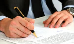 A man in a suit, perhaps a courtier from Quebec, signs a submission with a black and gold pen. His left hand rests on the paper, showing a silver watch, while his right hand writes among the printed text and signatures. 