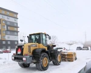 A yellow front loader equipped with a snow plough clears snow from a parking lot surrounded by snowdrifts. Cars are parked nearby and an apartment building stands on the left. Power lines are visible above, a sign that winter challenges will be met head-on.  