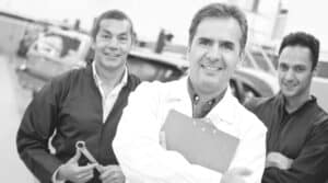 Three men stand in a garage. The man in front, in a white coat, acts like a courtier, paperweight in hand, and smiles confidently. Two men in overalls stand behind him, one holding a wrench. Blurry vehicles and tools fill the background.   