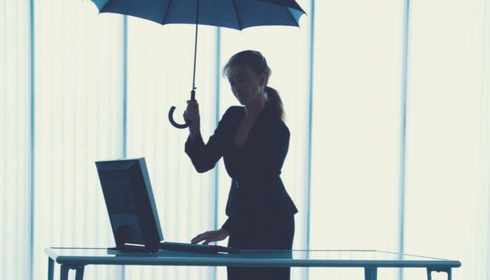 Une femme en tenue professionnelle se tient à un bureau avec un ordinateur, un parapluie ouvert à la main, comme si elle attendait un appel d'offres ou consultait un comparateur de prix. Le contre-jour dessine sa silhouette sur fond de grandes fenêtres aux stores verticaux.