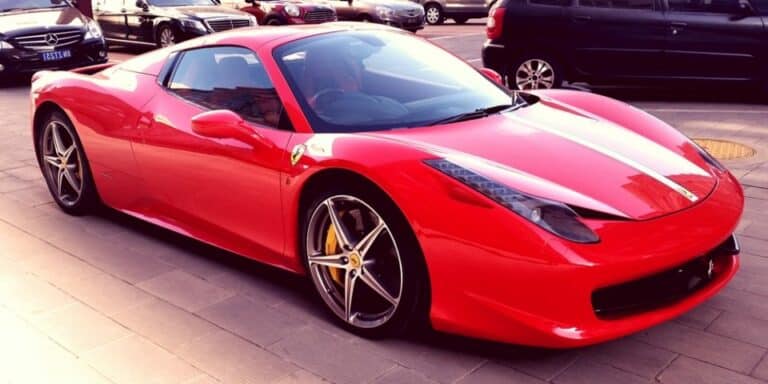 Une Ferrari rouge brillante aux accents noirs est garée sur une rue pavée, entourée d'autres voitures. Son design élégant et aérodynamique et ses étriers de frein dorés offrent une garantie visuelle de performance parmi une Mercedes-Benz et d'autres véhicules garés.