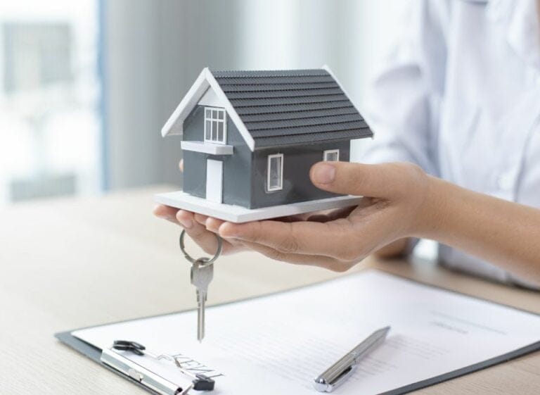 Une personne portant une chemise blanche tient une petite maison modèle et une clé – une image qui capture le processus immobilier au Québec, impliquant peut-être un courtisan préparant des documents et une soumission pour l'achat d'une maison sur une table en bois clair.