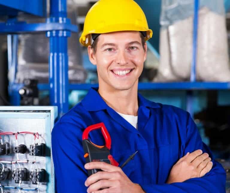 A smiling electrician, wearing a yellow helmet and blue overalls, stands with his arms crossed, a clamp meter in his hand. In this industrial environment with visible wiring, he offers expert service and a clear quote for each project.