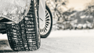 Gros plan sur un pneu arrière de voiture aux sculptures profondes roulant sur une route enneigée : idéal pour ceux qui recherchent la tranquillité d'esprit grâce à un comparateur d'assurance avant leurs trajets hivernaux. La neige s'accroche au pneu tandis que des arbres flous en hiver remplissent l'arrière-plan.