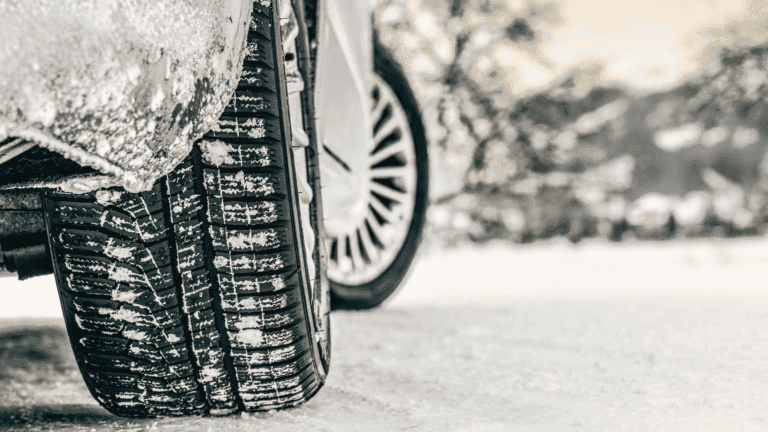 Gros plan sur un pneu arrière de voiture aux sculptures profondes roulant sur une route enneigée : idéal pour ceux qui recherchent la tranquillité d'esprit grâce à un comparateur d'assurance avant leurs trajets hivernaux. La neige s'accroche au pneu tandis que des arbres flous en hiver remplissent l'arrière-plan.