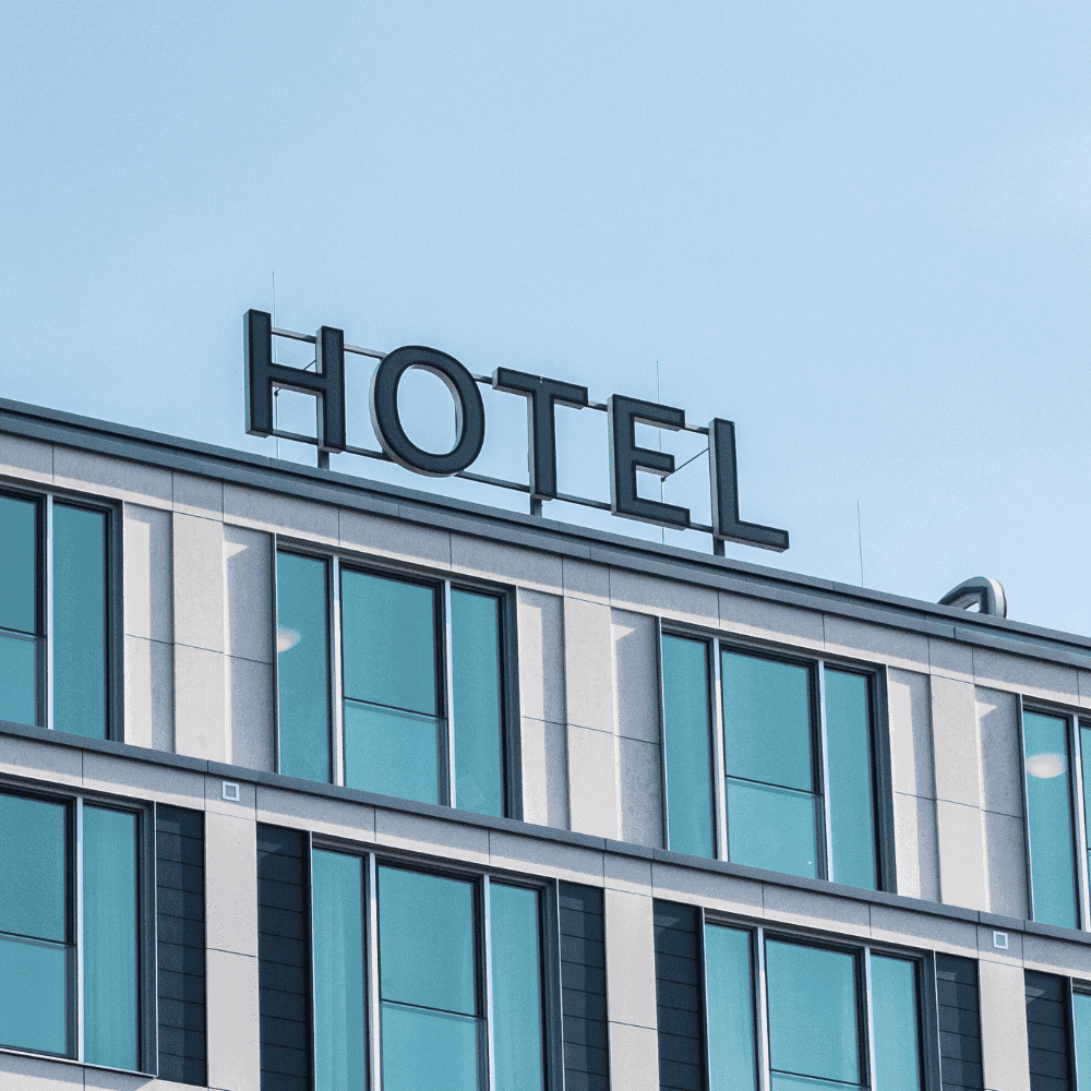 Un bâtiment moderne aux grandes fenêtres teintées de bleu et au toit plat arbore un panneau « HOTEL » audacieux sur un ciel dégagé : idéal pour ceux qui recherchent l'Assurance Hôtels ou une offre personnalisée. Sa façade contemporaine se distingue par ses lignes épurées.