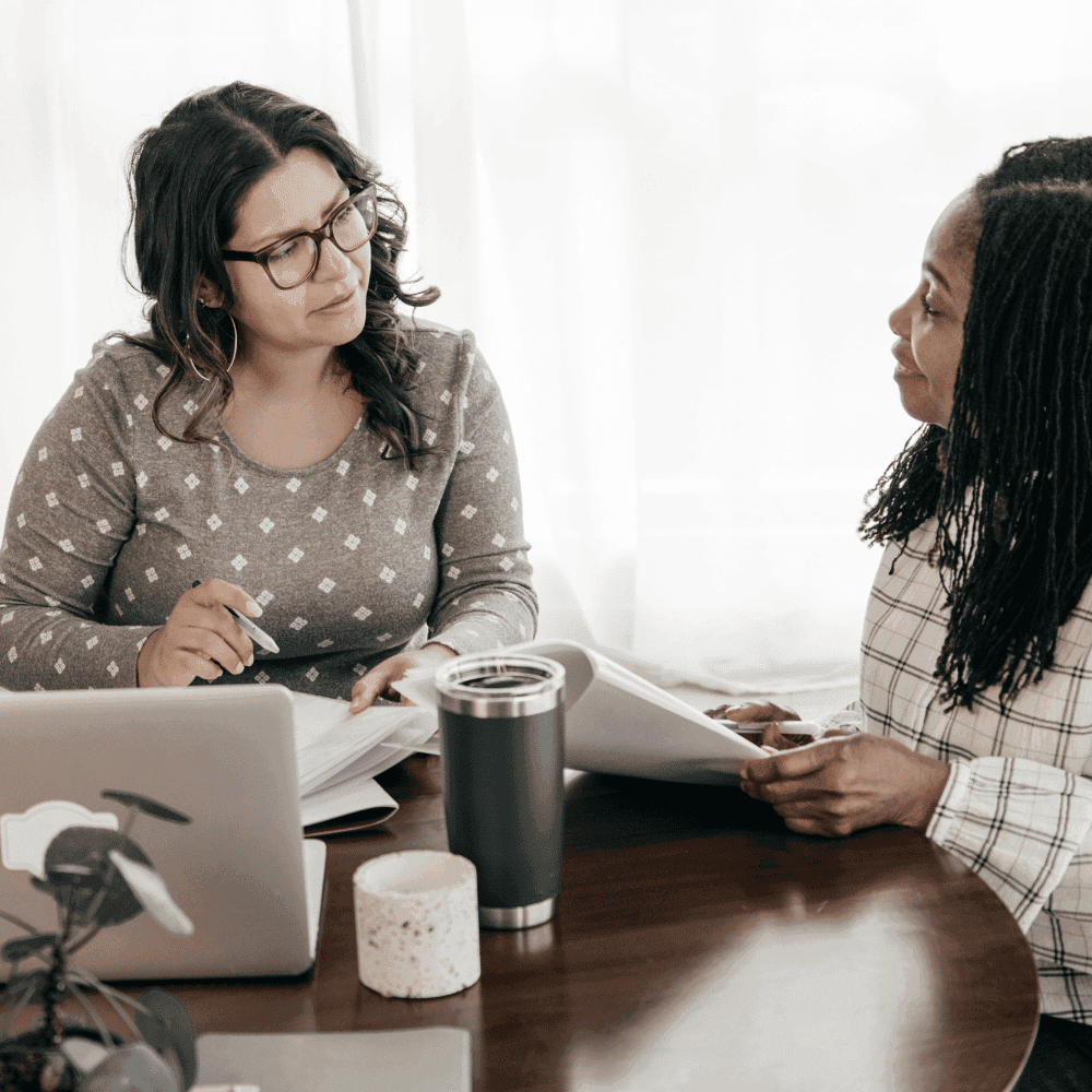 Deux femmes sont assises à une table ronde et discutent de formalités administratives liées aux assurances et aux erreurs et omissions. L'une porte des lunettes et un haut gris à motifs, tient un stylo et écoute attentivement ; un ordinateur portable, un verre et une plante sont posés sur la table devant elles.