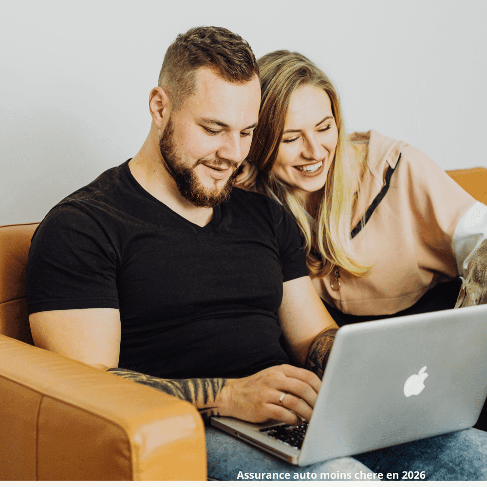 A smiling man and woman sit next to each other on a brown couch in front of a laptop screen. The man types while the woman leans over the screen, both looking happy and engaged as they search for car insurance options on their Apple laptops 