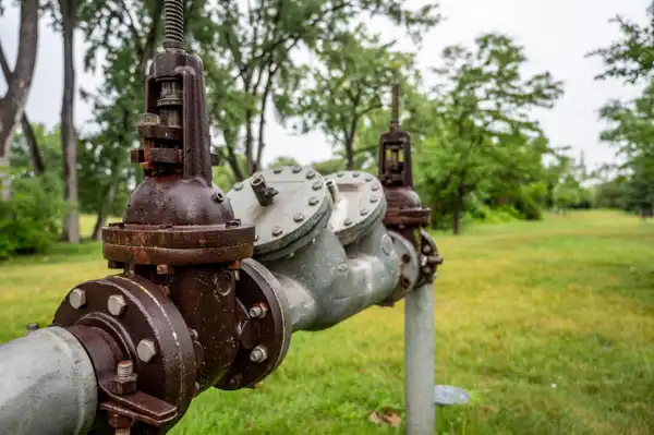 Metal pipe with valves and rust in a grassy area with trees in the background.