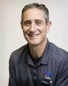 A person with short hair smiles warmly, wearing a dark collared shirt featuring the "About Us" logo. The backdrop is plain and light-colored, highlighting their friendly demeanor.