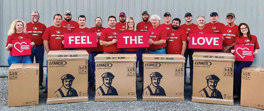 Group of people in matching red shirts holding "Feel the Love" signs, standing behind large Lennox boxes in front of a gray building.