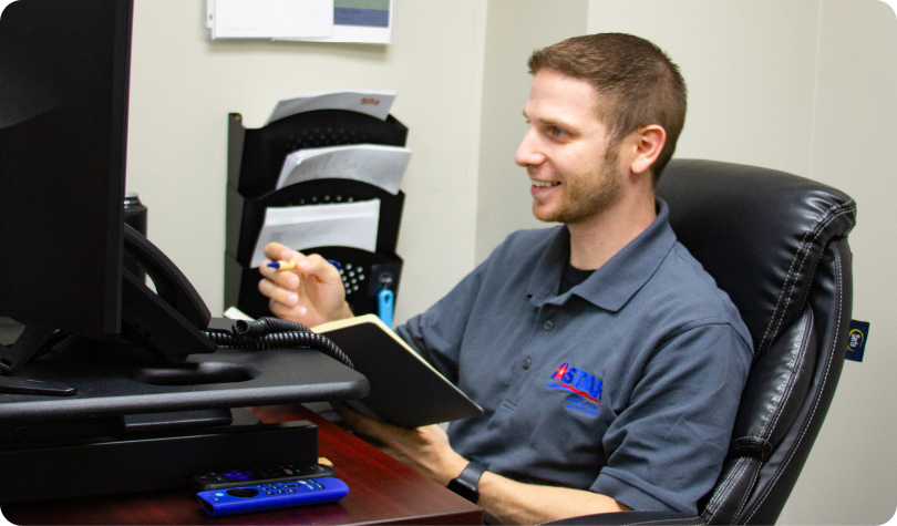 A man in a gray polo sits at a desk, holding a notebook and pen, intently studying an Astar interface on the computer screen. A remote control is on the desk.