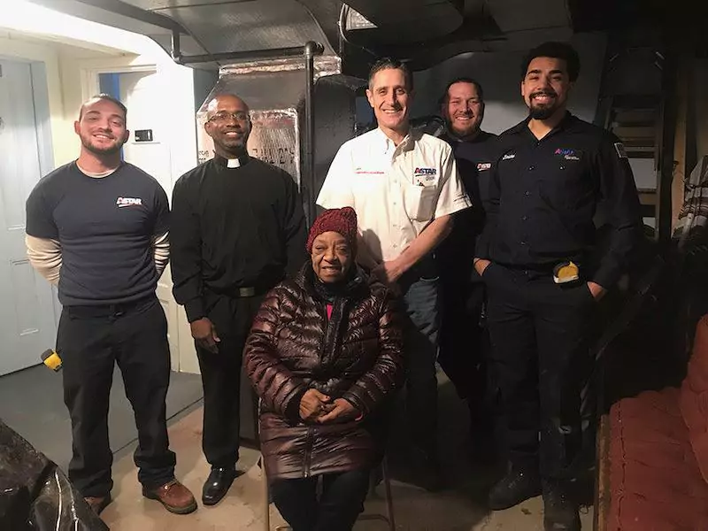A seated woman in a red coat is surrounded by five standing men in work uniforms and a clergy outfit, posing for a photo indoors.
