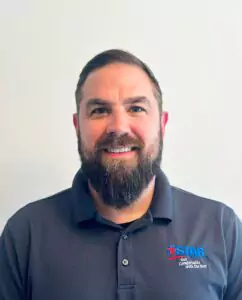 A man with a beard, wearing a dark polo shirt adorned with the company logo, smiles at the camera against a plain background, embodying the welcoming spirit of our "About Us" story.