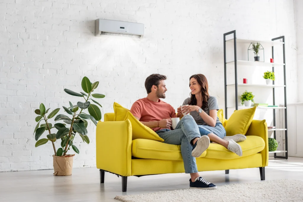 A couple sits on a yellow sofa in a modern living room, holding mugs, with plants and shelves in the background. An air conditioner is visible on the white brick wall.