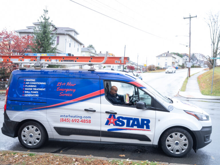 A blue and white Astar service van is parked on a suburban street with a person seated inside; a ladder is secured on the van's roof.