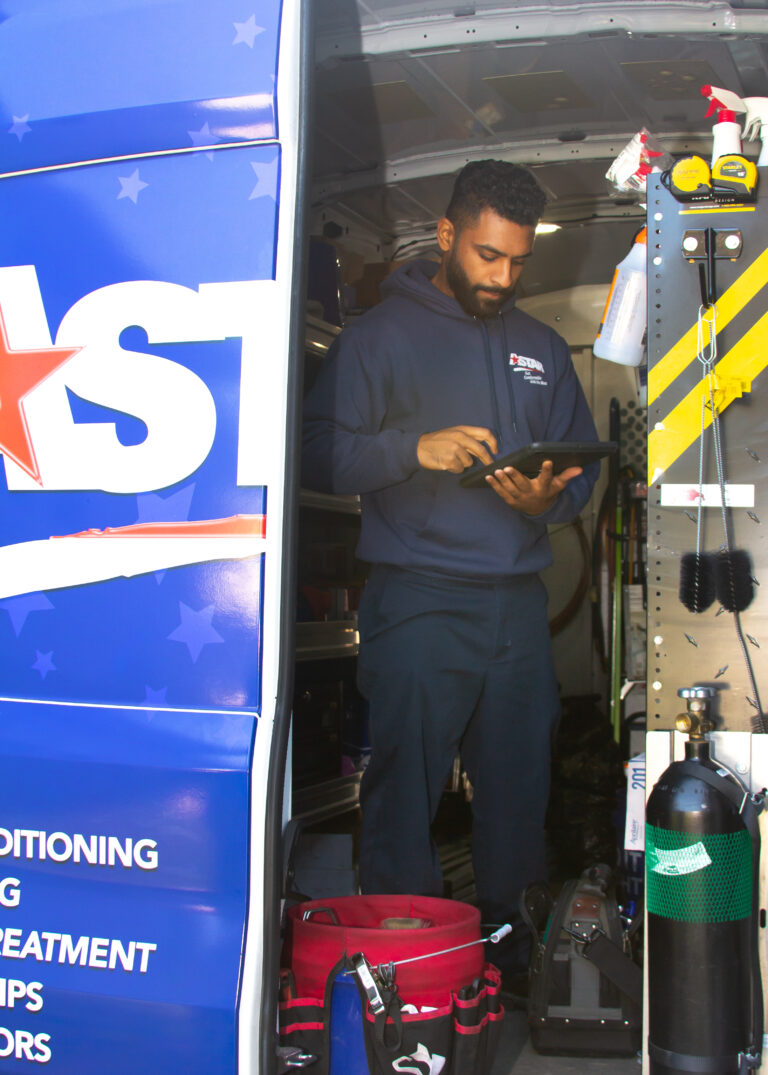 A technician in a navy hoodie uses a tablet while standing inside a service van equipped with tools and equipment.