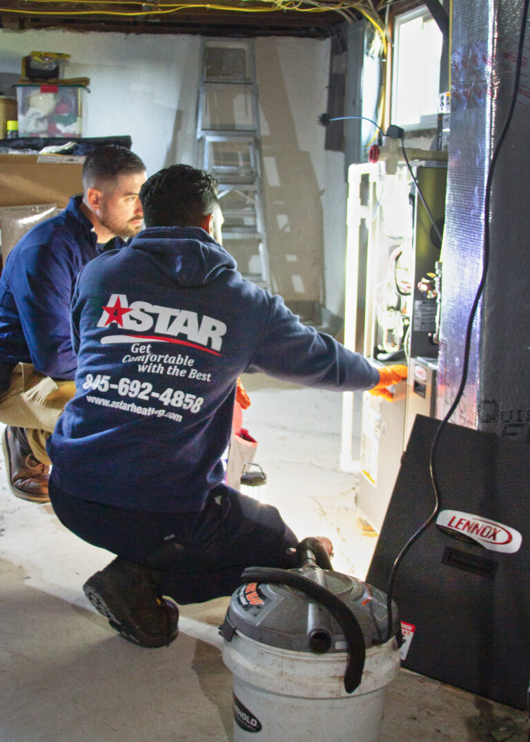 Two technicians work on a Lennox HVAC unit in a basement. One wears a "Astar" sweatshirt and the other observes. Tools and a vacuum are nearby.