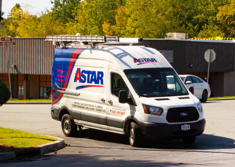 A white Astar service van with a ladder on top is parked on a street near a commercial building and trees.