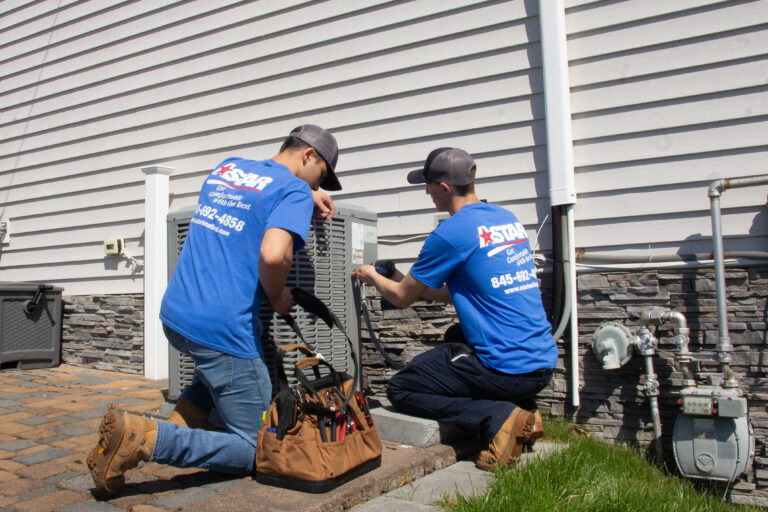 Two HVAC technicians in blue uniforms work on an outdoor air conditioning unit beside a house, with tools and equipment nearby.