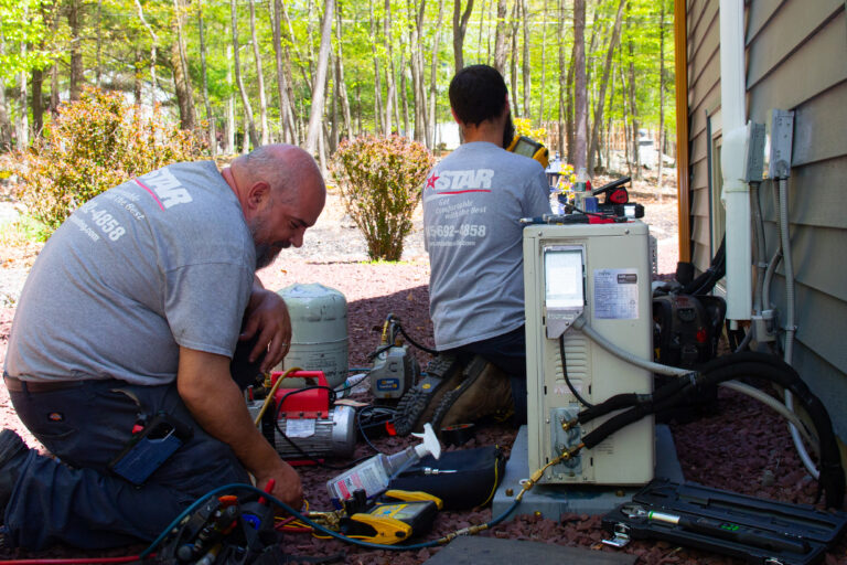 Two HVAC technicians in gray shirts repair or inspect an outdoor air conditioning unit next to a house on a sunny day.