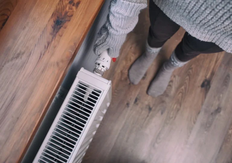 Person wearing gray gloves and sweater adjusts a thermostat on a white radiator, standing on a wooden floor.