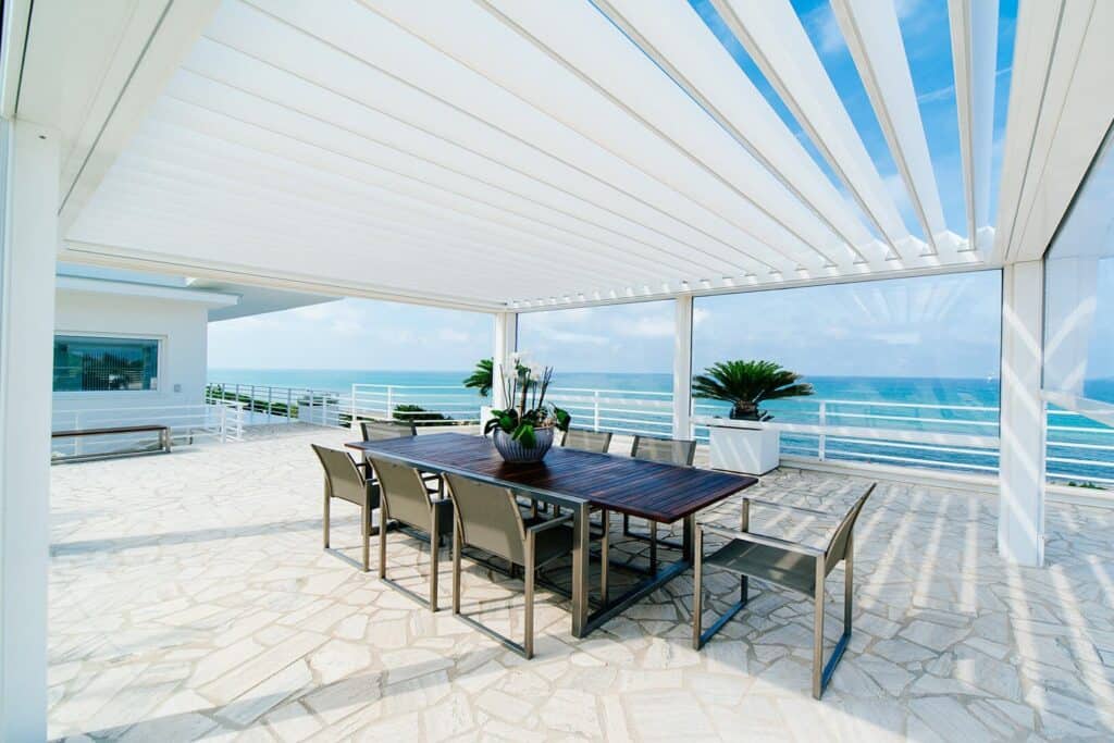 Soggiorno all'aperto con vista mare, copertura in lamelle automatiche, spazio pranzo elegante e piante tropicali, ideale per relax e convivialità durante le calde giornate estive.