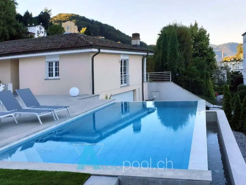 Piscina interrata a sfioro con acqua cristallina, estesa sul terrazzo di una casa privata in collina, circondata da vegetazione e con sedie per il relax.