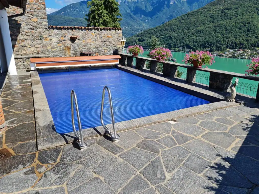 Piscina interrata con copertura blu, situata in un ambiente con vista sui monti e lago, decorata con fiori colorati sulla balaustra.