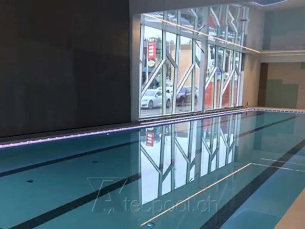 Piscina coperta a sfioro con pareti in vetro e illuminazione LED, ambiente moderno e elegante.
