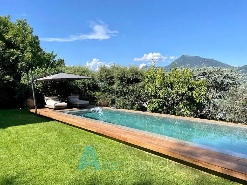 Piscina interrata a sfioro con deck in legno e area relax con sedie e ombrellone, immersa nel verde con vista sulle montagne.
