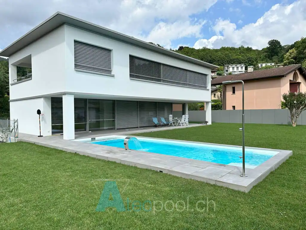 Piscina interrata a sfioro con bordo in pietra, situata in un giardino ben curato con una moderna casa bianca sullo sfondo.