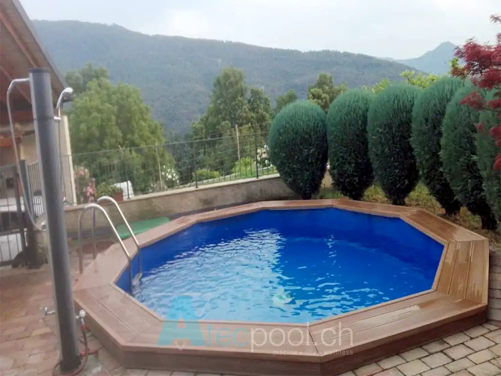 Piscina interrata a sfioro con bordo in legno e contornata da arbusti sempreverdi, immersa in un paesaggio naturale con montagne sullo sfondo.