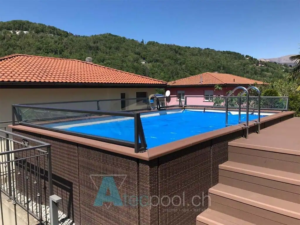 Piscina interrata con copertura a tettoia, immersa nel contesto di case con tetti rossi e natura montana sullo sfondo.