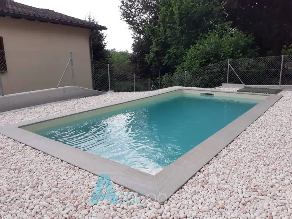 Azzurro cristallino di una piscina interrata con bordo in cemento, circondata da ghiaia bianca sotto un cielo nuvoloso.
