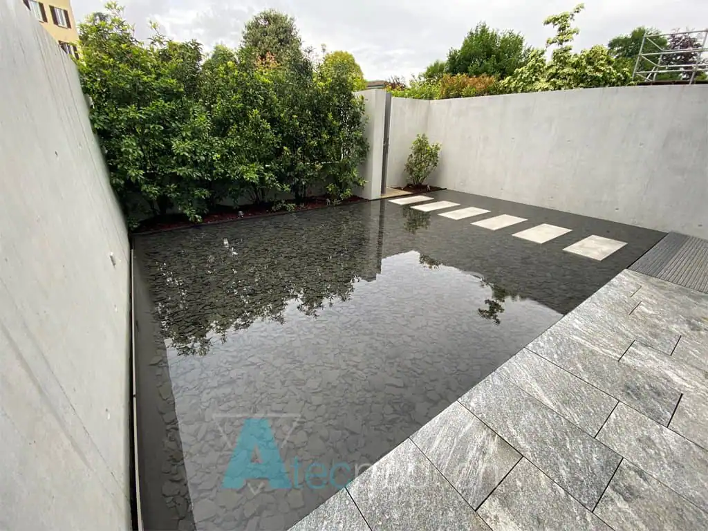 Piscina interrata con pavimentazione in pietra e bordi in cemento, circondata da vegetazione e muri di sicurezza in cemento, perfetta per il relax e il benessere.