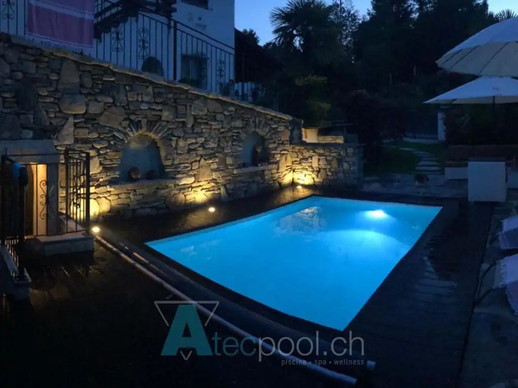 Piscina interrata con illuminazione di notte, rivestita in mosaico blu e circondata da pietre naturali e luci soffuse.