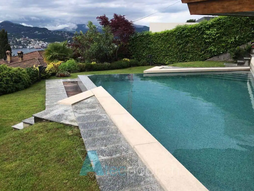Piscina interrata con bordo a sfioro in un giardino ben curato con vista panoramica sulla città e il lago.