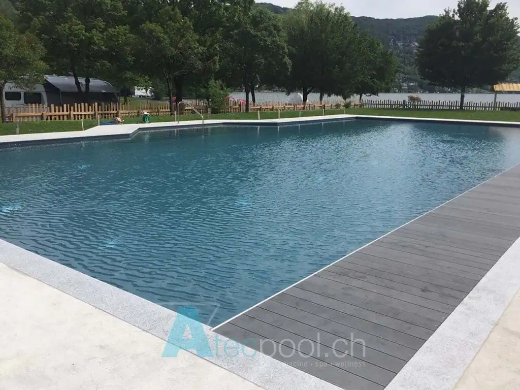 Piscina interrata con rivestimento in liner blu e bordo in cemento, immersa nel verde di un giardino con alberi e vista sul lago. Ideale per relax e attività sportive all'aperto.