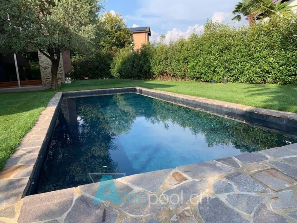 Piscina interrata con bordo in pietra, immersa in un giardino verde e ombreggiata dagli alberi, perfetta per il relax e il nuoto all'aperto.