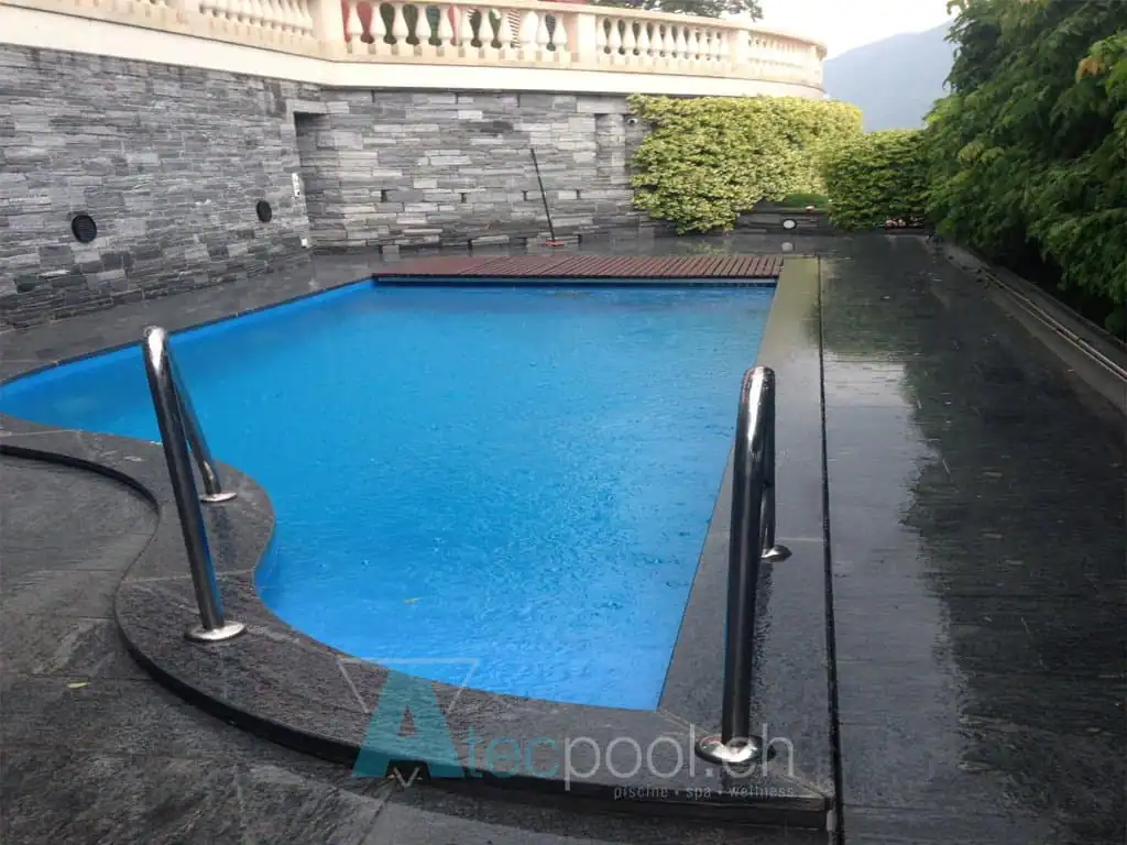 Piscina interrata con bordo a sfioro, rivestimento in piastrelle nere e scalettine in acciaio. Ambiente esterno con piante e pareti in pietra, ideale per relax e wellness.