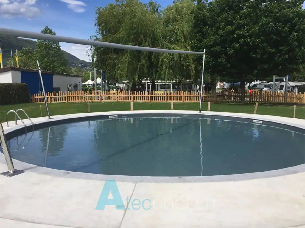 Piscina interrata rotonda con bordo bianco e scaletta in acciaio, immersa nel verde di un'area dedicata al relax e al benessere.