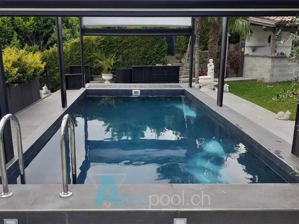 Piscina interrata a sfioro in giardino, copertura in vetro, con zona relax e piante ornamentali.