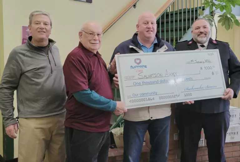 Four men proudly stand before an indoor staircase, holding a large $1,000 check made out to the Salvation Army, embodying our "about us" commitment to giving back.