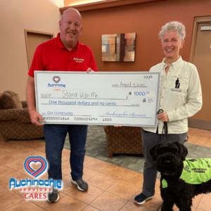 Two people proudly hold a $1,000 check from Auchinachie Services, highlighting the company's commitment to community support as seen in their 'About Us' values. Meanwhile, a black dog wearing a service vest stands loyally at the forefront.