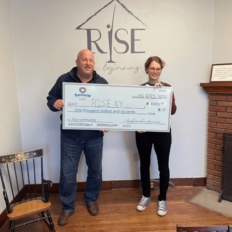 Two people hold a large ceremonial check for $1000 from Automate Care to RISENY, embodying the spirit of our "About Us" values, as they stand in a cozy room with a fireplace and chairs.